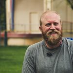 A cheerful bearded man smiling outdoors in a casual setting with greenery.