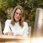 A woman in glasses writing and working on her laptop outdoors in natural daylight.