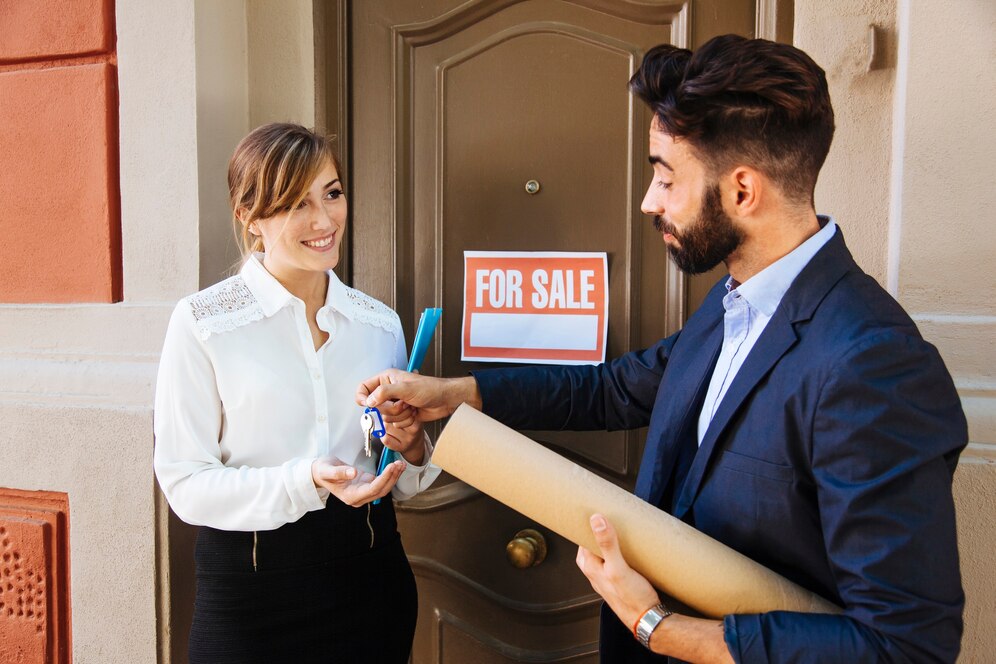 man and woman stand in front of a house displaying a for sale sign, smiling and looking at the camera